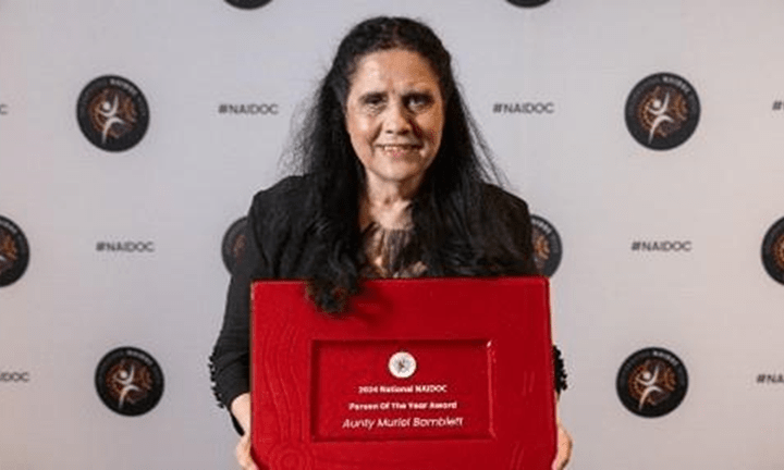 Photo of Aunty Muriel Bamblett AM standing looking directly at the camera holding an award